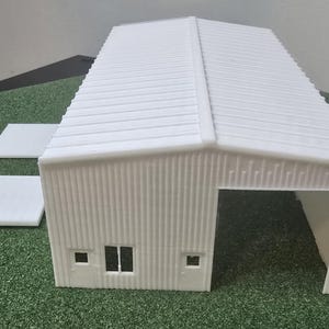 May include: A white model of a corrugated metal building with a gabled roof. The building has a large opening on one side and two small windows. Two white rectangular pieces are next to the building.