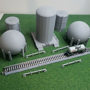 May include: A collection of gray miniature industrial models, including cylindrical tanks, spherical tanks, and a rail car with the text "BUDIUM". The models are arranged on a green surface with a section of model train tracks.