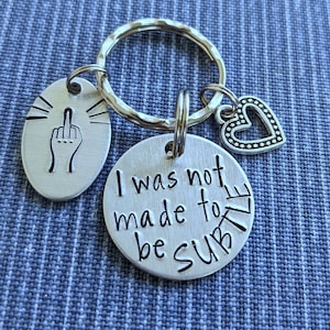 May include: Silver keychain with a middle finger charm, a heart charm, and a round charm that says "I was not made to be subtle".
