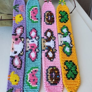 Op de afbeelding: Vier kleurrijke vriendschapsarmbanden met verschillende pixelontwerpen. De armbanden zijn gemaakt van kleurrijk borduurgaren en hebben een kuiken, een watermeloen, een donut en een paddenstoel.