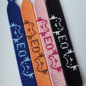 Puede incluir: Cuatro pulseras de la amistad con diferentes hilos de colores y palabras tejidas en el diseño. Las pulseras son azules, naranjas, rosas y negras. Las palabras en las pulseras son "Believe", "Awesome", "Happy" y "Friendship".