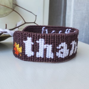 Puede incluir: Pulsera de amistad marrón con letras blancas que dicen "gracias" y un pequeño diseño de hoja naranja, amarilla y blanca.