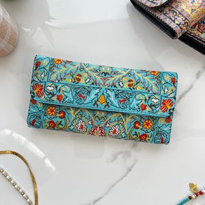 May include: A turquoise clutch wallet with a detailed floral pattern in red, yellow, and white. The wallet is rectangular with a fold-over flap. The design features stylized flowers and leaves, creating a vintage aesthetic.