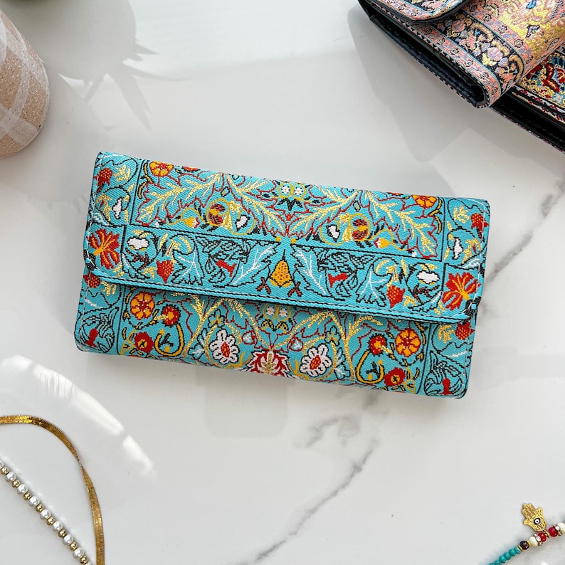 May include: A turquoise clutch wallet with a detailed floral pattern in red, yellow, and white. The wallet is rectangular with a fold-over flap. The design features stylized flowers and leaves, creating a vintage aesthetic.