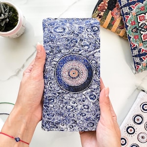 Puede incluir: Una cartera rectangular de tela azul texturizada con un prominente motivo circular de ojo turco en el centro. El dise&ntilde;o incluye patrones intrincados de c&iacute;rculos conc&eacute;ntricos en tonos azul oscuro, azul claro y blanco, creando un efecto visual detallado.