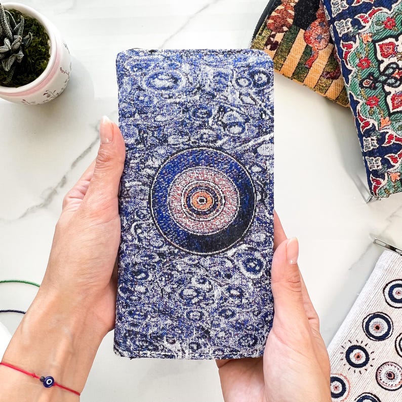 Puede incluir: Una cartera rectangular de tela azul texturizada con un prominente motivo circular de ojo turco en el centro. El dise&ntilde;o incluye patrones intrincados de c&iacute;rculos conc&eacute;ntricos en tonos azul oscuro, azul claro y blanco, creando un efecto visual detallado.