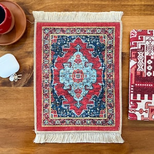 Puede incluir: Una alfombrilla rectangular con un patrón central de medallón en azul claro y rojo, rodeado por un borde en azul oscuro y rojo con flecos de color crema en los bordes superior e inferior, con motivos geométricos y florales detallados.