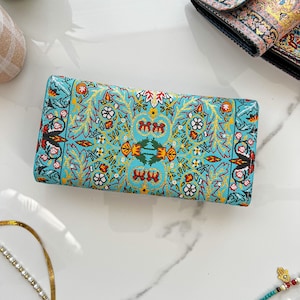 May include: A rectangular turquoise wallet with an intricate floral pattern in gold, red, and white. The wallet features a detailed design with symmetrical floral and leaf motifs. The overall aesthetic is ornate and decorative.
