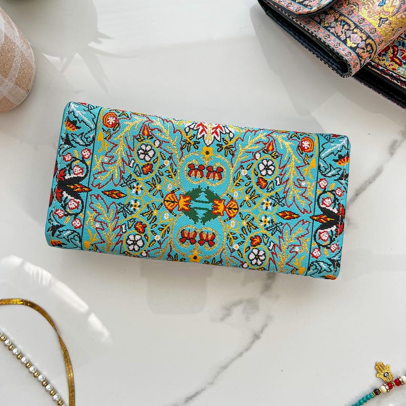 May include: A rectangular turquoise wallet with an intricate floral pattern in gold, red, and white. The wallet features a detailed design with symmetrical floral and leaf motifs. The overall aesthetic is ornate and decorative.