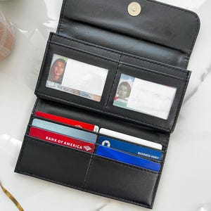 May include: Black bi-fold wallet with multiple card slots and clear ID windows. The wallet is open, revealing credit cards and identification cards. The wallet is made of a smooth, matte material and has a snap closure.