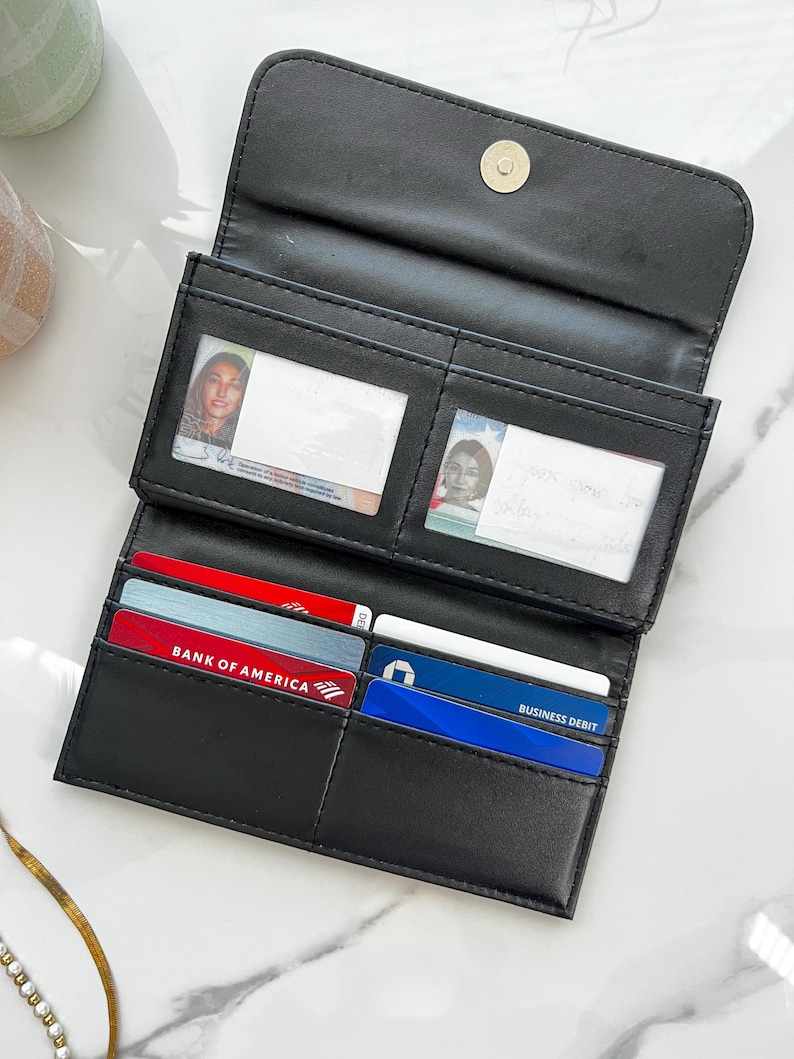 May include: Black bi-fold wallet with multiple card slots and clear ID windows. The wallet is open, revealing credit cards and identification cards. The wallet is made of a smooth, matte material and has a snap closure.