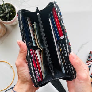 Puede incluir: Una cartera rectangular con cremallera y un exterior de tela con estampado multicolor. El interior cuenta con m&uacute;ltiples ranuras para tarjetas, un compartimento central con cremallera y bolsillos abiertos que contienen varias tarjetas de cr&eacute;dito, identificaciones y billetes.