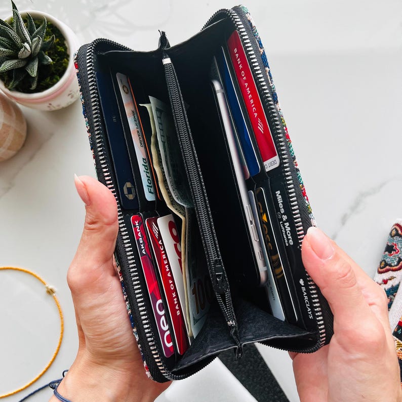 Puede incluir: Una cartera rectangular con cremallera y un exterior de tela con estampado multicolor. El interior cuenta con m&uacute;ltiples ranuras para tarjetas, un compartimento central con cremallera y bolsillos abiertos que contienen varias tarjetas de cr&eacute;dito, identificaciones y billetes.