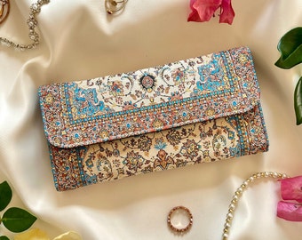 May include: A rectangular wallet with a detailed, colorful pattern in shades of beige, blue, and red. The wallet is closed and lies on a white satin surface, surrounded by jewelry and flowers. The design resembles a traditional rug.
