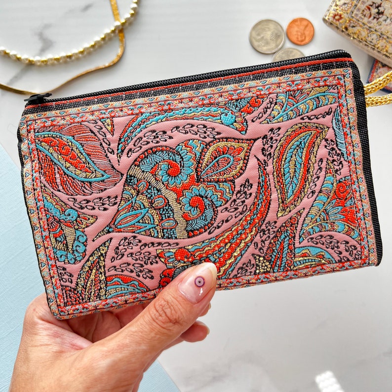 May include: A pink fabric coin purse with a colorful paisley design. The purse has a black zipper and a black trim.