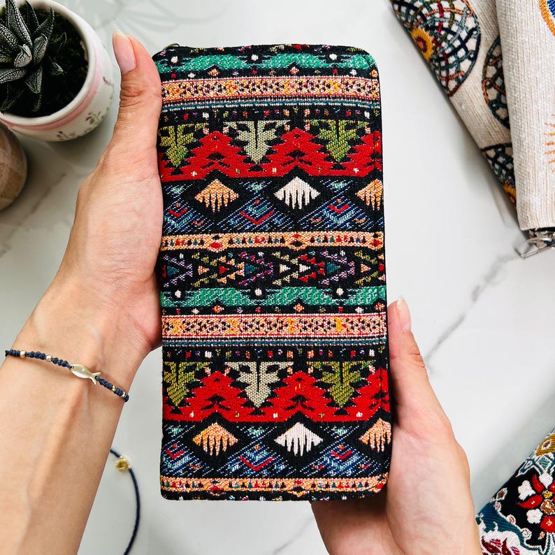 Puede incluir: Una cartera rectangular con un exterior de tela tejida con un patr&oacute;n geom&eacute;trico repetitivo en negro, rojo, verde, naranja y blanco. El dise&ntilde;o incluye bandas horizontales de formas estilizadas y l&iacute;neas dentadas en toda la superficie.