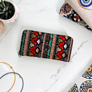 Puede incluir: Una cartera rectangular con cremallera que presenta un vibrante patr&oacute;n de tapiz tejido con formas geom&eacute;tricas en rojo, negro, azul, verde y amarillo. La cartera tiene un tirador de cremallera de metal plateado y un exterior de tela estructurada.