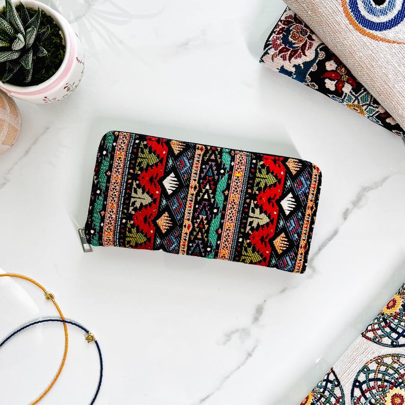 Puede incluir: Una cartera rectangular con cremallera que presenta un vibrante patr&oacute;n de tapiz tejido con formas geom&eacute;tricas en rojo, negro, azul, verde y amarillo. La cartera tiene un tirador de cremallera de metal plateado y un exterior de tela estructurada.