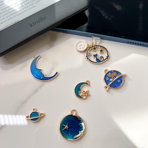 May include: A set of six gold-toned charms with blue and green enamel. The charms feature a crescent moon, a star, a planet, a cat, and a moon with a star. The charms are perfect for adding a touch of whimsy to jewelry or crafts.