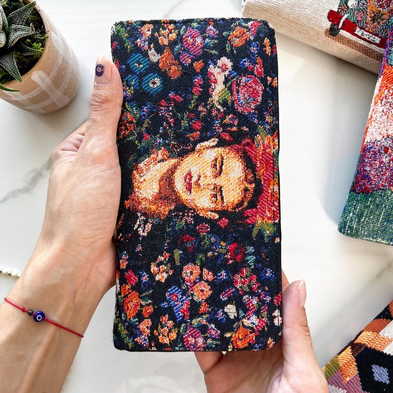 Puede incluir: Una cartera rectangular con un dise&ntilde;o de tapiz tejido sobre un fondo negro. El patr&oacute;n muestra un retrato central de una persona rodeado por una densa disposici&oacute;n de flores coloridas en tonos rojos, naranjas, azules y verdes.