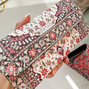 May include: A patterned clutch with a fold-over flap. The design features a repeating floral pattern in red, black, and cream. The clutch is rectangular and appears to be made of a woven fabric.