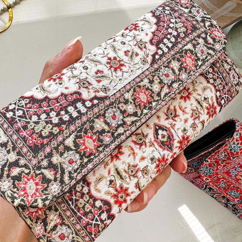 May include: A patterned clutch with a fold-over flap. The design features a repeating floral pattern in red, black, and cream. The clutch is rectangular and appears to be made of a woven fabric.