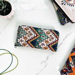 Puede incluir: Una cartera rectangular con cremallera que presenta un exterior de tela tejida con un patr&oacute;n geom&eacute;trico multicolor en tonos turquesa, naranja, blanco y azul oscuro. La cartera tiene un cierre de cremallera negro en el borde superior.
