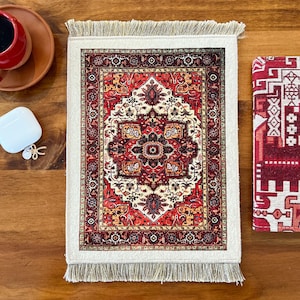 May include: A rectangular fabric mat featuring a detailed red, cream, and black ornate pattern with a central medallion and intricate border designs. The mat has light-colored fringe trim on the top and bottom edges, creating a textured finish.