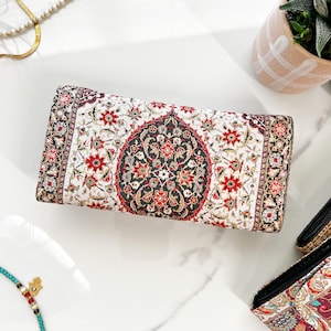 May include: A rectangular clutch with an intricate floral pattern in red, black, and gold on a white background. The design resembles a traditional rug. The clutch is likely made of fabric and has a rectangular shape.
