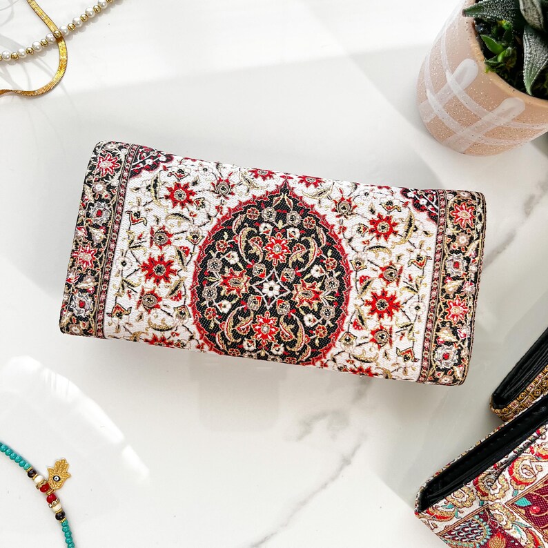 May include: A rectangular clutch with an intricate floral pattern in red, black, and gold on a white background. The design resembles a traditional rug. The clutch is likely made of fabric and has a rectangular shape.