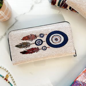 Puede incluir: Una cartera rectangular de color beige con textura tejida, que presenta un s&iacute;mbolo de ojo turco azul oscuro y tres plumas colgantes en tonos rojo, marr&oacute;n y verde. La cartera tiene un cierre de cremallera negro en la parte superior y lateral.