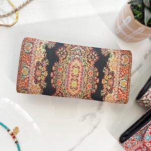 May include: A rectangular clutch with a detailed, colorful pattern. The design features a dark background with red, yellow, and green floral motifs. The edges are trimmed with a similar patterned border. The clutch is lying on a white surface.