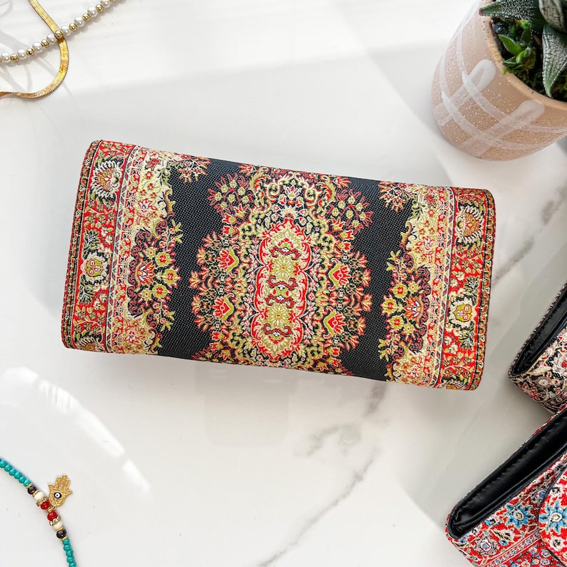 May include: A rectangular clutch with a detailed, colorful pattern. The design features a dark background with red, yellow, and green floral motifs. The edges are trimmed with a similar patterned border. The clutch is lying on a white surface.