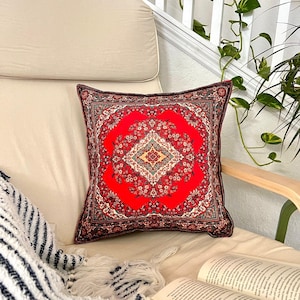 Könnte beinhalten: Ein rotes und schwarzes Kissen mit einem Muster, das einem persischen Teppich ähnelt. Das Kissen liegt auf einem beigen Sofa mit einer gestreiften Decke und einem Buch.