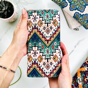 Puede incluir: Una cartera rectangular con un exterior de tela tejida con un patr&oacute;n geom&eacute;trico repetitivo en tonos azul verdoso, azul oscuro, rojo y dorado sobre un fondo blanco roto. El dise&ntilde;o incluye l&iacute;neas en zigzag, diamantes y peque&ntilde;os motivos circulares.
