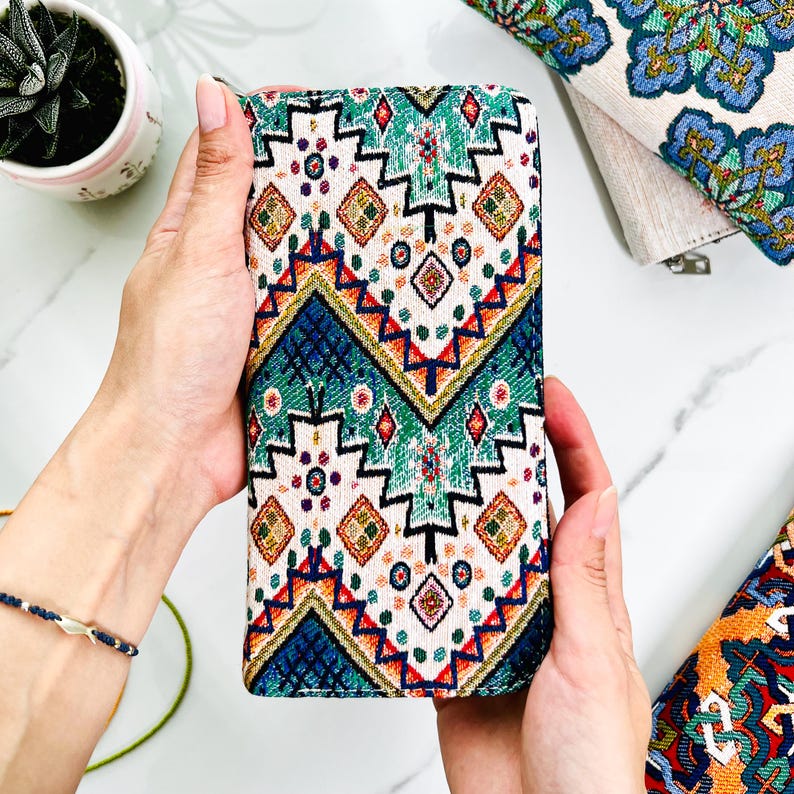 Puede incluir: Una cartera rectangular con un exterior de tela tejida con un patr&oacute;n geom&eacute;trico repetitivo en tonos azul verdoso, azul oscuro, rojo y dorado sobre un fondo blanco roto. El dise&ntilde;o incluye l&iacute;neas en zigzag, diamantes y peque&ntilde;os motivos circulares.
