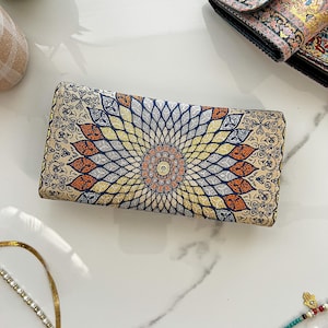May include: A rectangular clutch with an intricate floral pattern in beige, orange, yellow, and blue. The design radiates from a central medallion. The clutch is likely made of fabric and has a decorative, ornate style.