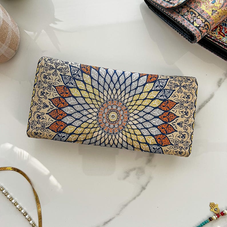 May include: A rectangular clutch with an intricate floral pattern in beige, orange, yellow, and blue. The design radiates from a central medallion. The clutch is likely made of fabric and has a decorative, ornate style.