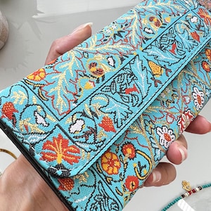 May include: A turquoise clutch with an embroidered floral pattern in red, gold, and white. The design features stylized flowers and leaves, creating a vibrant and detailed aesthetic. The clutch is held open, showcasing the interior.