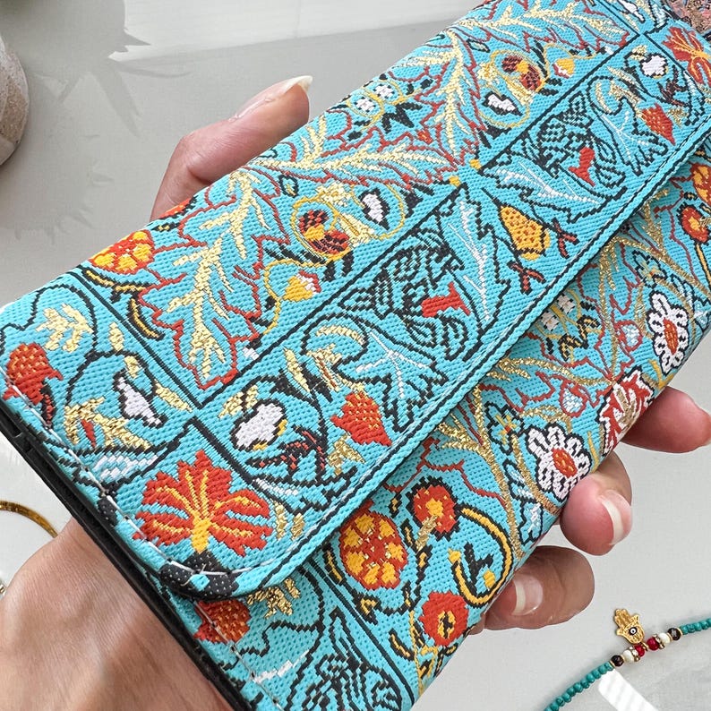 May include: A turquoise clutch with an embroidered floral pattern in red, gold, and white. The design features stylized flowers and leaves, creating a vibrant and detailed aesthetic. The clutch is held open, showcasing the interior.