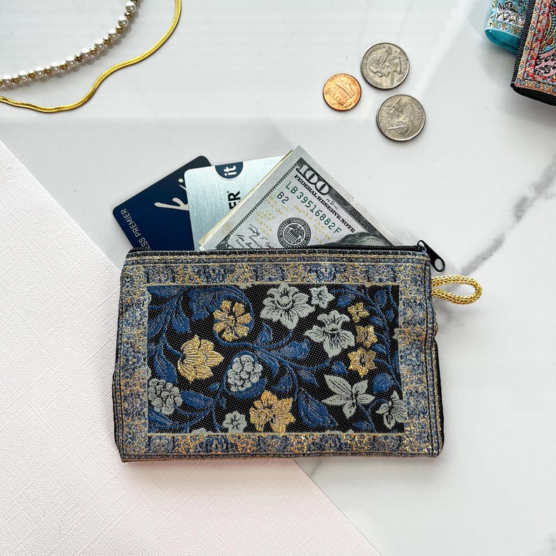 May include: A small black zippered pouch with a gold zipper pull. The pouch is decorated with a floral pattern in shades of blue, gold, and white. The pouch is open and contains a credit card, a dollar bill, and some coins.