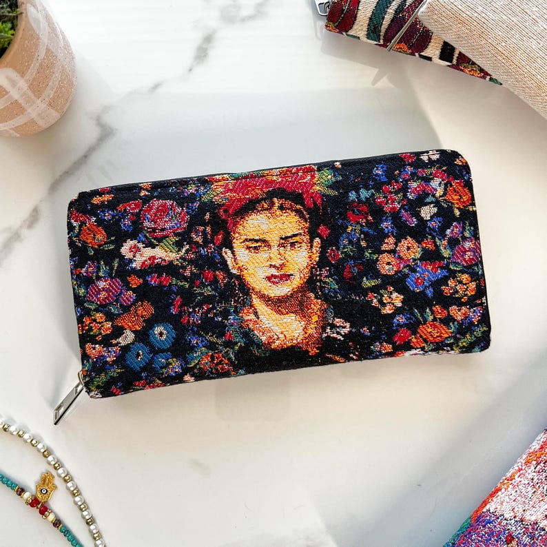 Puede incluir: Una cartera rectangular con cremallera que presenta un dise&ntilde;o de tapiz tejido de Frida Kahlo rodeada por un vibrante patr&oacute;n floral en tonos rojos, azules, naranjas y verdes sobre un fondo negro, con un tirador de cremallera met&aacute;lico plateado.
