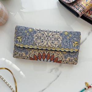May include: A rectangular clutch with a detailed floral pattern in blue, beige, and orange. The clutch has a fold-over flap with gold trim and a decorative design. The background is a white marble surface.