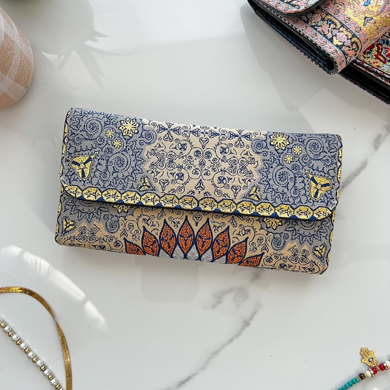 May include: A rectangular clutch with a detailed floral pattern in blue, beige, and orange. The clutch has a fold-over flap with gold trim and a decorative design. The background is a white marble surface.