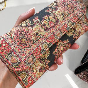 May include: A rectangular clutch with a detailed floral pattern in red, gold, and green on a black background. The clutch has a fold-over flap and appears to be made of a woven fabric. The design resembles a traditional rug.