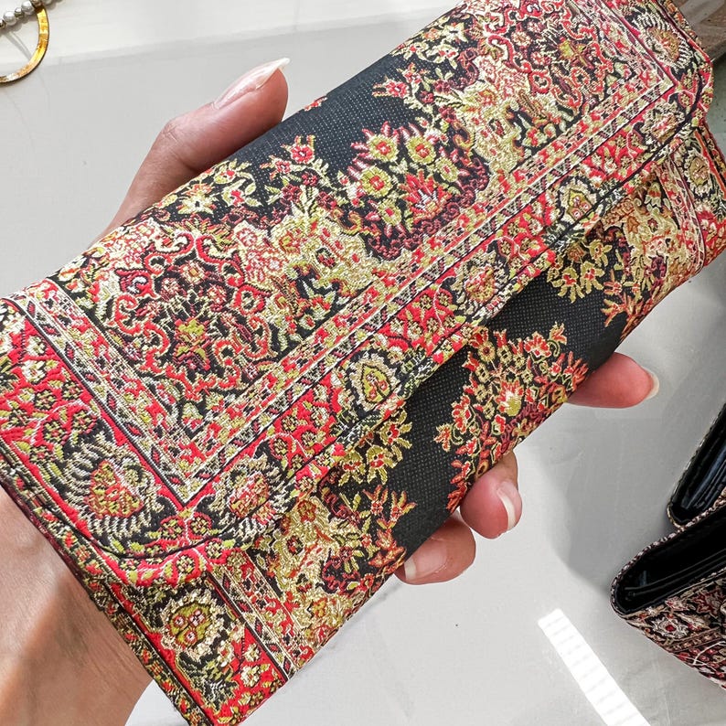 May include: A rectangular clutch with a detailed floral pattern in red, gold, and green on a black background. The clutch has a fold-over flap and appears to be made of a woven fabric. The design resembles a traditional rug.