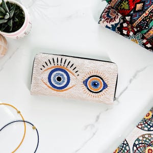 Puede incluir: Una cartera rectangular con base de tela texturizada en color blanco roto que presenta dos motivos de ojos bordados. Los ojos est&aacute;n representados en tonos azul, negro y naranja con l&iacute;neas radiantes. La cartera tiene una cremallera negra.