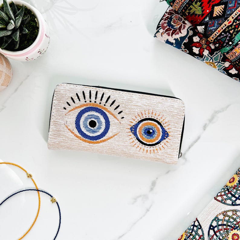 Puede incluir: Una cartera rectangular con base de tela texturizada en color blanco roto que presenta dos motivos de ojos bordados. Los ojos est&aacute;n representados en tonos azul, negro y naranja con l&iacute;neas radiantes. La cartera tiene una cremallera negra.