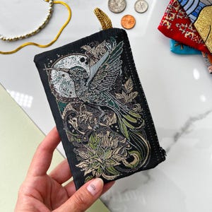 May include: A black fabric pouch with a zipper closure features an embroidered design of a green and silver bird with a long tail, surrounded by floral elements. The pouch has a dark background with a subtle shimmer.
