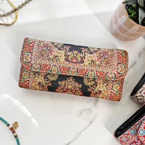 May include: A rectangular clutch with a black background and a detailed red, yellow, and green floral pattern. The clutch has a fold-over flap and appears to be made of a woven fabric. The design resembles a traditional rug.